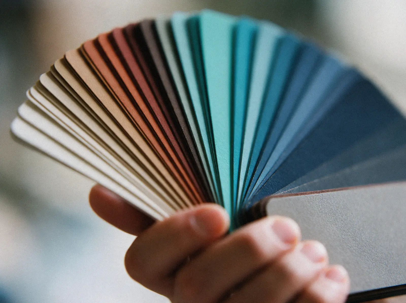 Eine Hand fächert einen Farbfächer auf, die Streifen laufen von Naturtönen über Türkis bis Blau