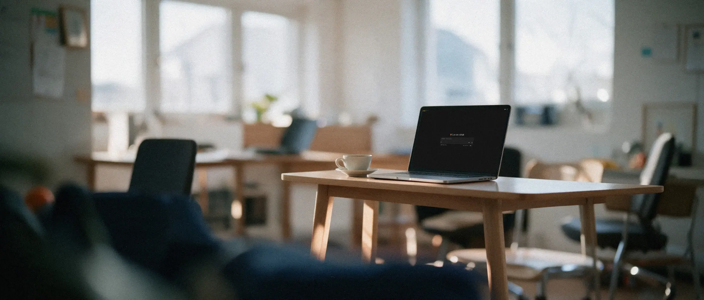 Leeres, helles Büro am frühen Morgen, auf einem Tisch ein aufgeklappter Laptop mit einem KI-Assistenten und eine Tasse