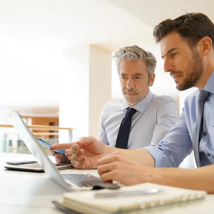 2 Männer die am Tischen sitzen und auf ein Macbook schauen